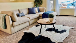 Una sala de estar con un sofá seccional color beige, una mesa redonda de centro con una planta, y un tapete de piel de vaca blanco y negro.