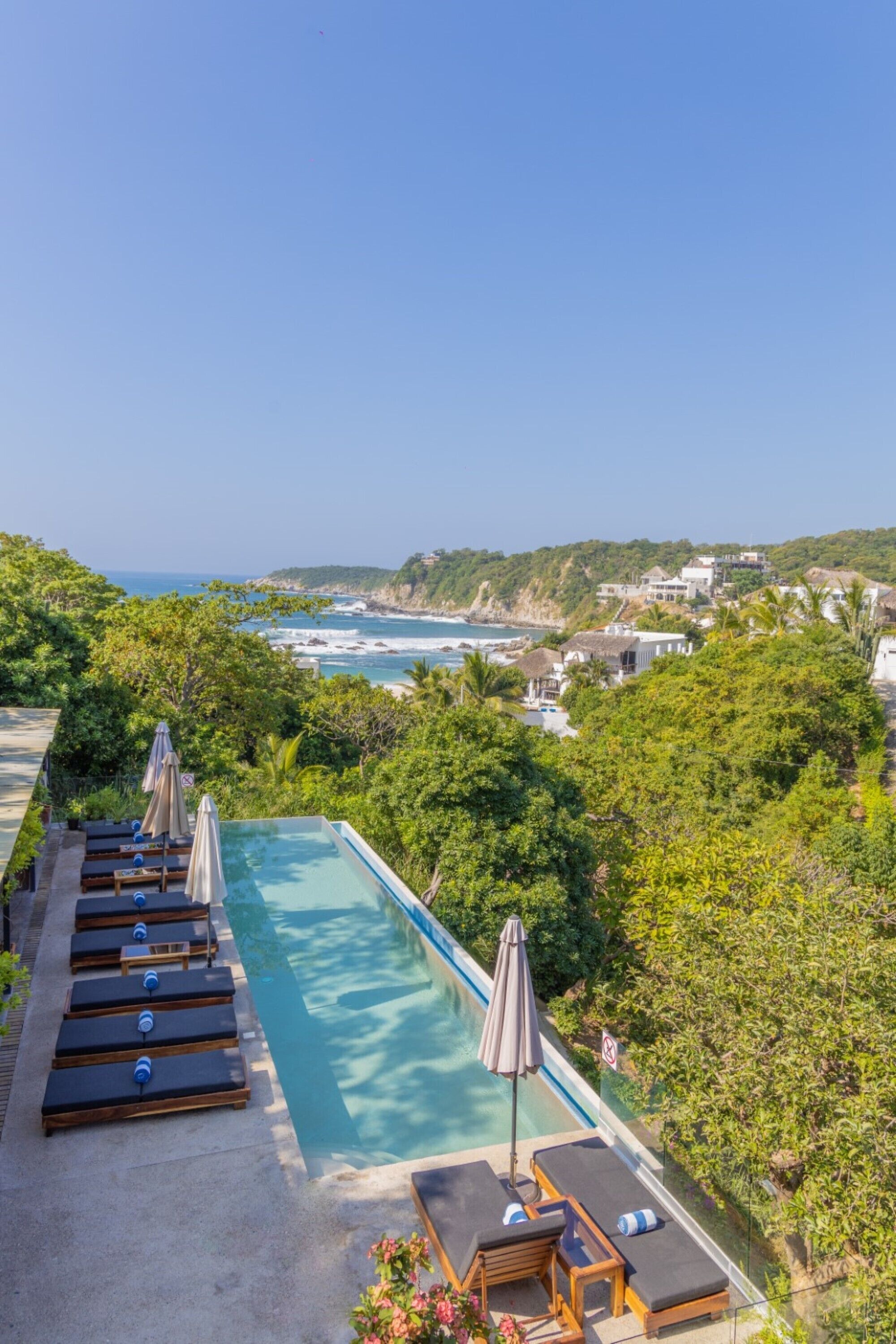 A rooftop infinity pool with lounge chairs and umbrellas, overlooking a coastal landscape with houses and greenery.