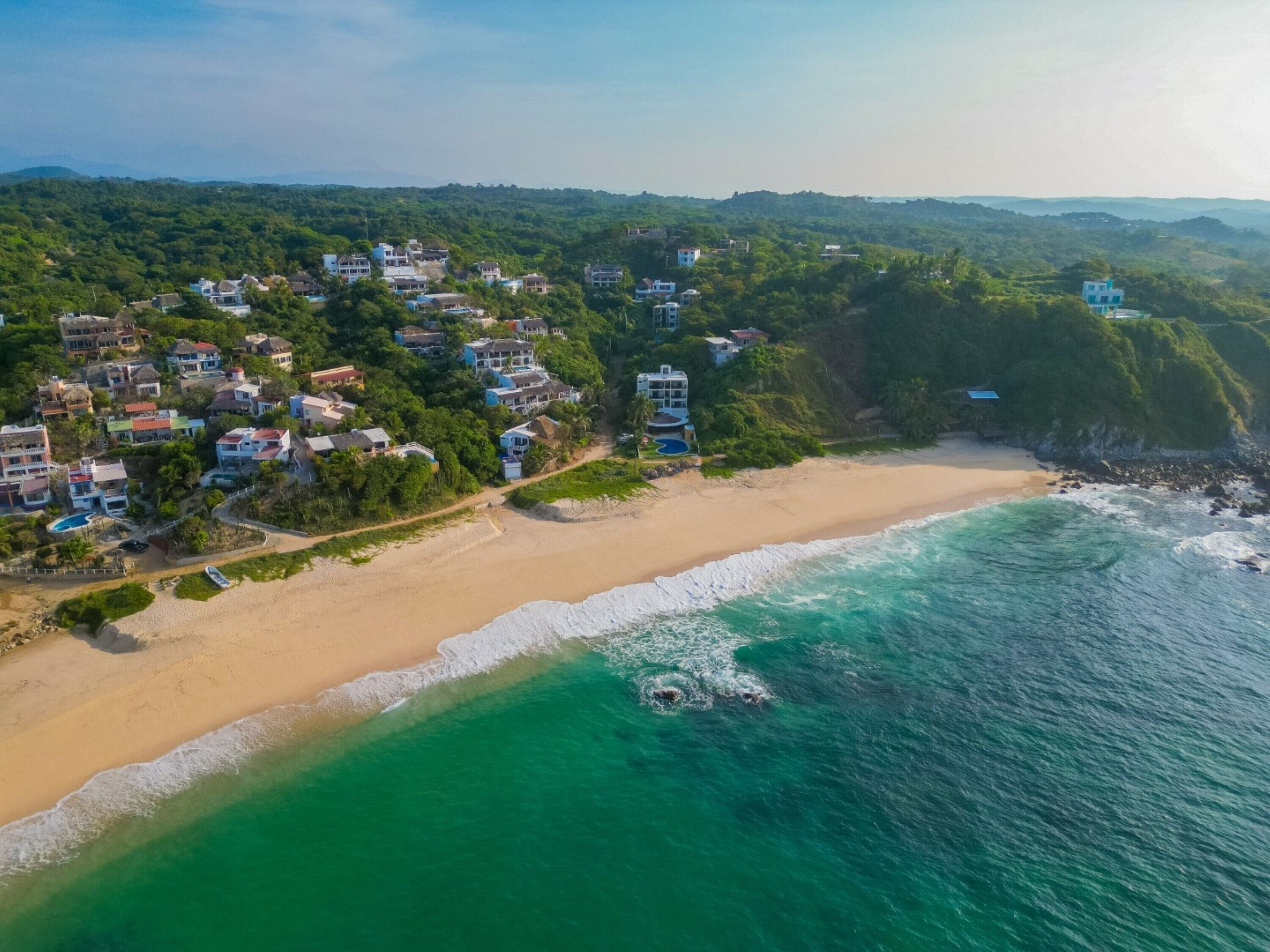 Vista aérea de un pueblo costero con una playa de arena, aguas turquesas cristalinas y un conjunto denso de edificios con diversos estilos arquitectónicos.