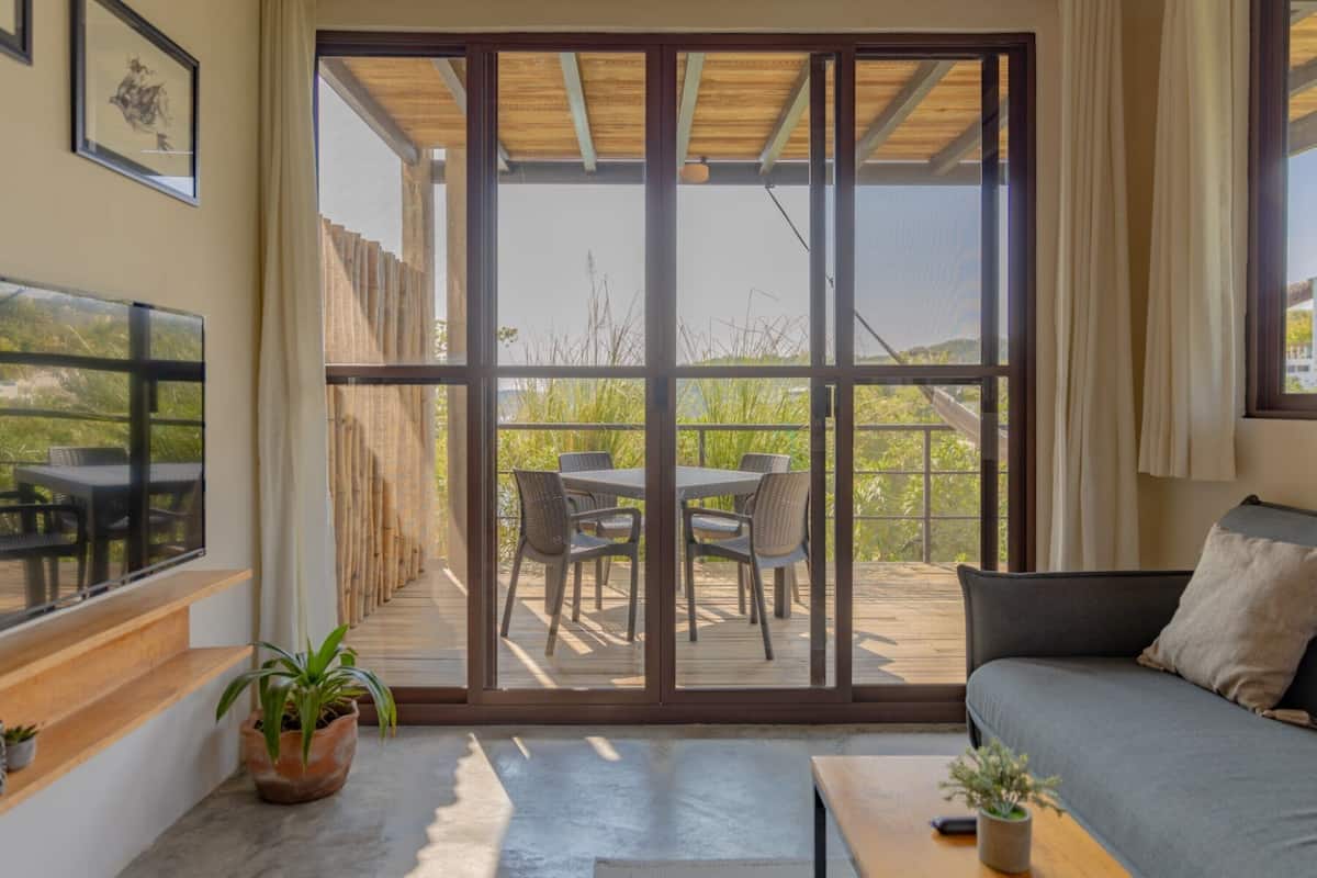 Una habitación con una puerta corrediza de vidrio que da a una terraza exterior con una mesa, sillas, un sofá y una pequeña mesa con una planta.