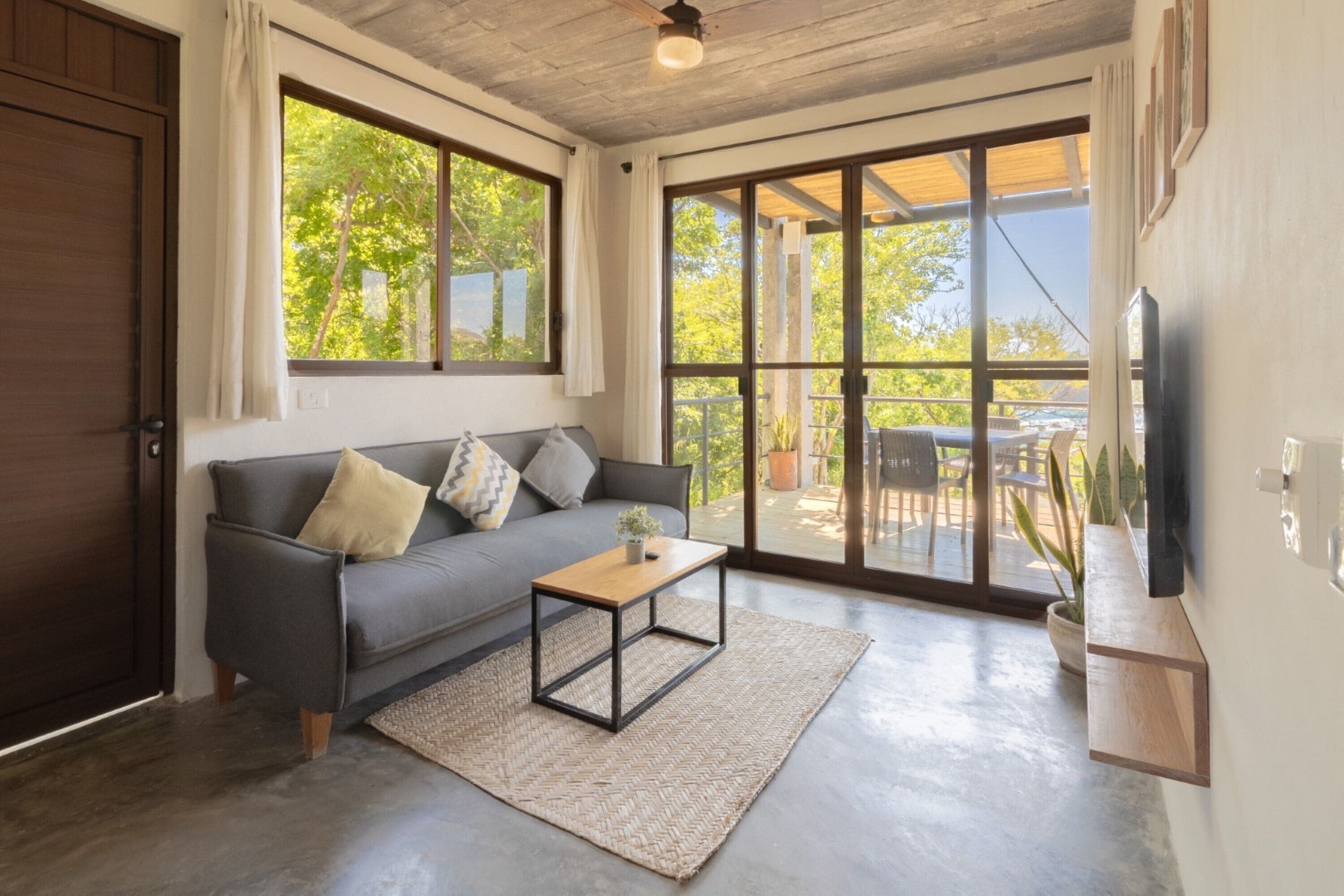 Un salon moderne avec un canapé gris, une table basse en bois et de grandes portes-fenêtres coulissantes donnant sur une terrasse extérieure.