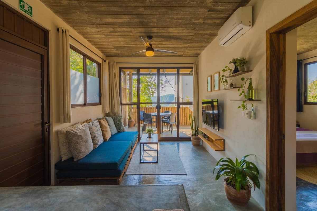 Una habitación con un sofá azul, una mesita, un ventilador de techo y un televisor en la pared.