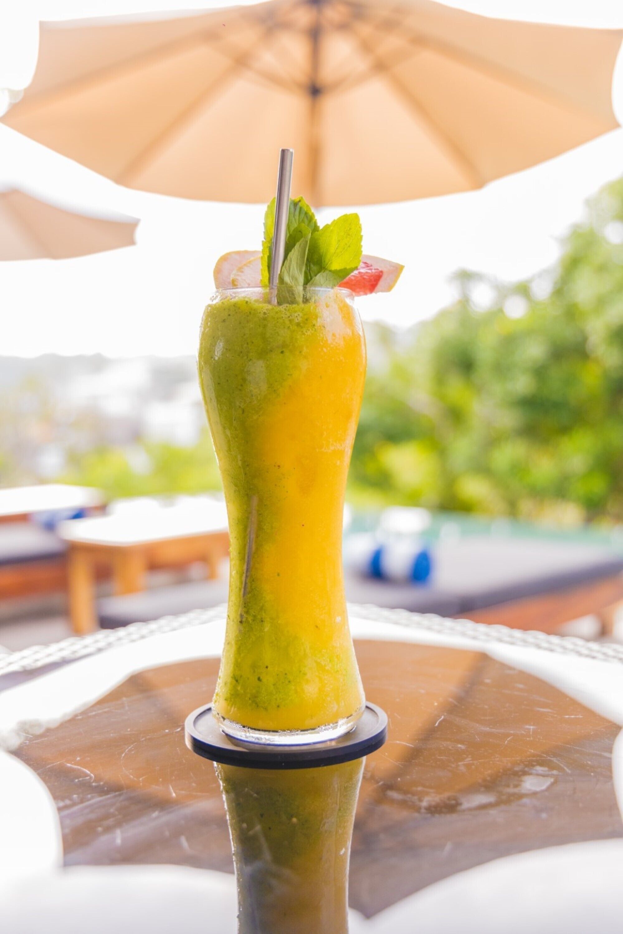 A tall glass with a green drink, garnished with mint and fruit, on a glass table with a metal base.
