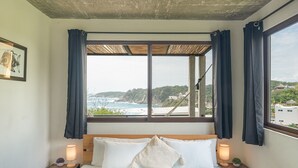 Une chambre avec une grande fenêtre donnant sur la côte, un lit avec du linge de lit blanc et deux lampes de chevet.