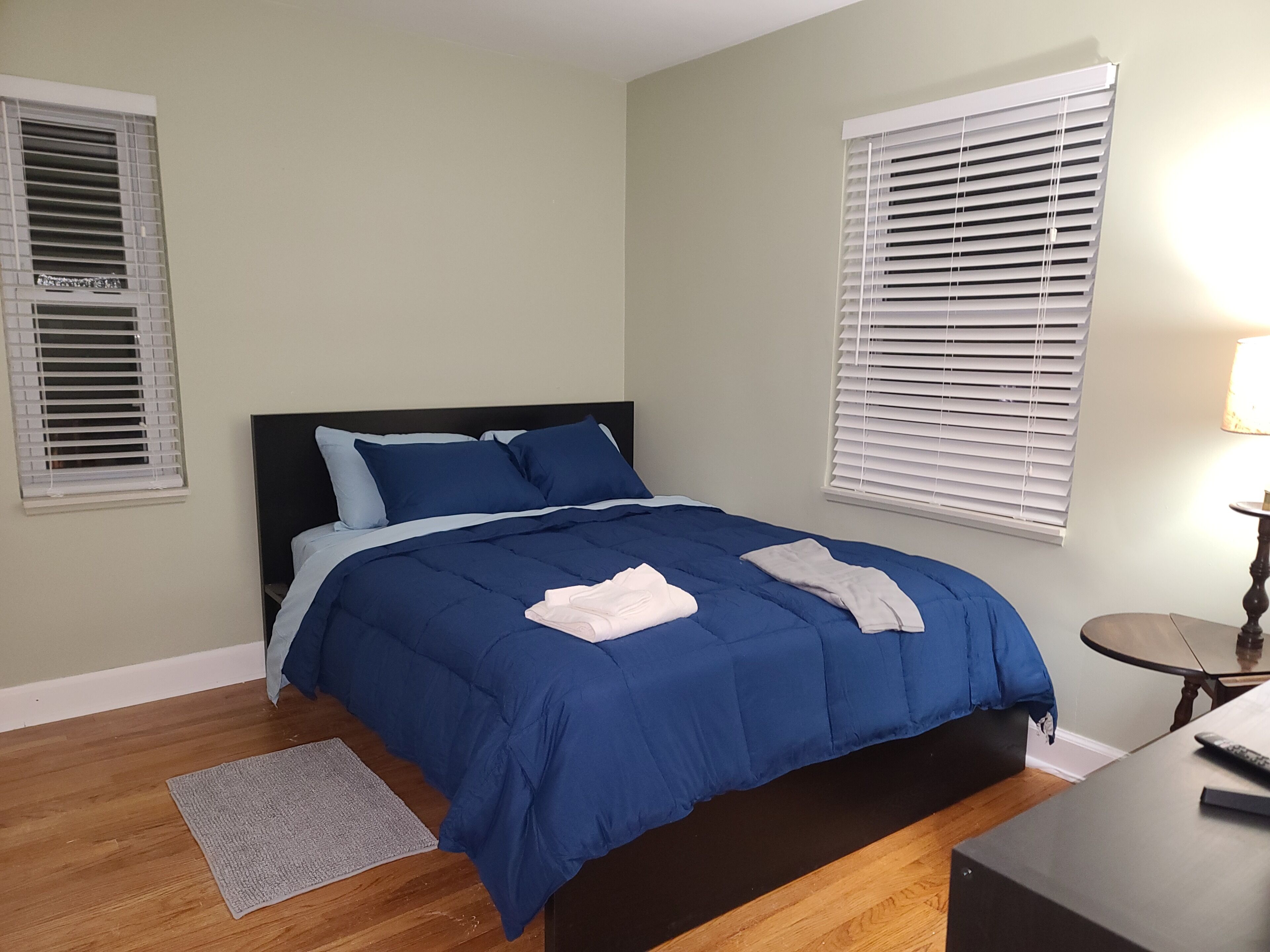 A bedroom with a bed, two windows with blinds, a bedside table, and a lamp.