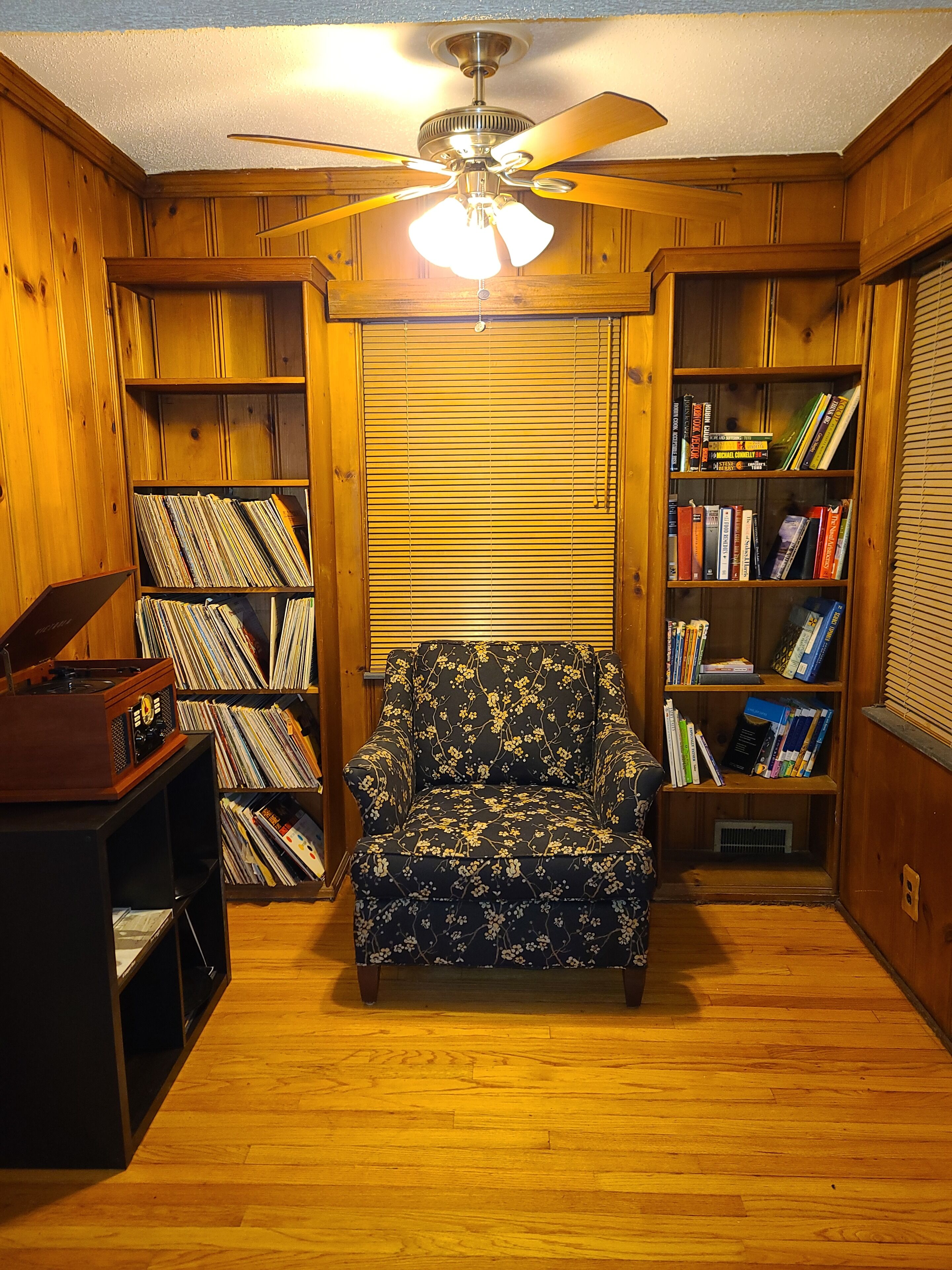 Une pièce chaleureuse avec des murs en bois, un ventilateur de plafond, une bibliothèque remplie de livres, un tourne-disque et un fauteuil à motifs floraux.