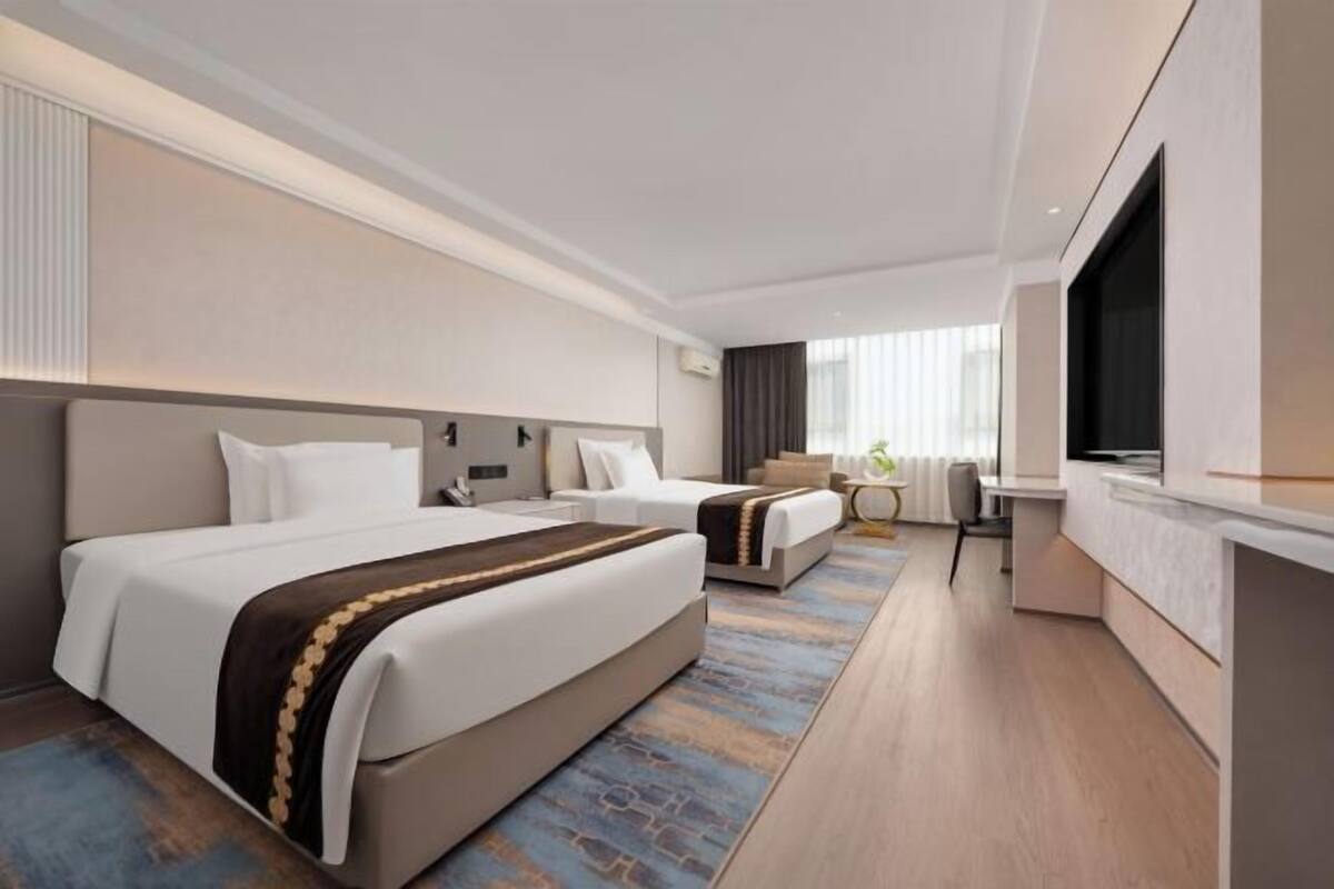 A modern hotel room with two beds, a TV, a desk, and a view of the city.