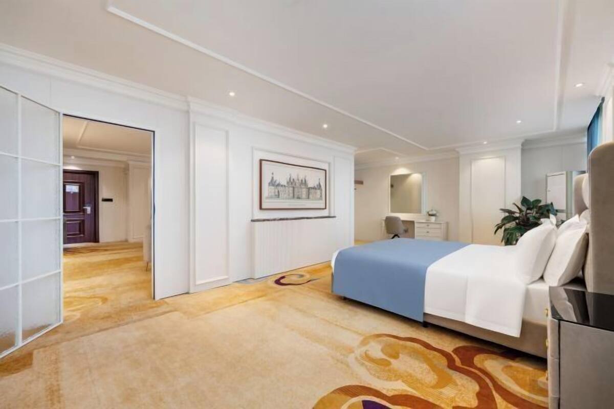 A hotel room with a bed, a desk, a chair, a mirror, a plant, and a framed picture on the wall.