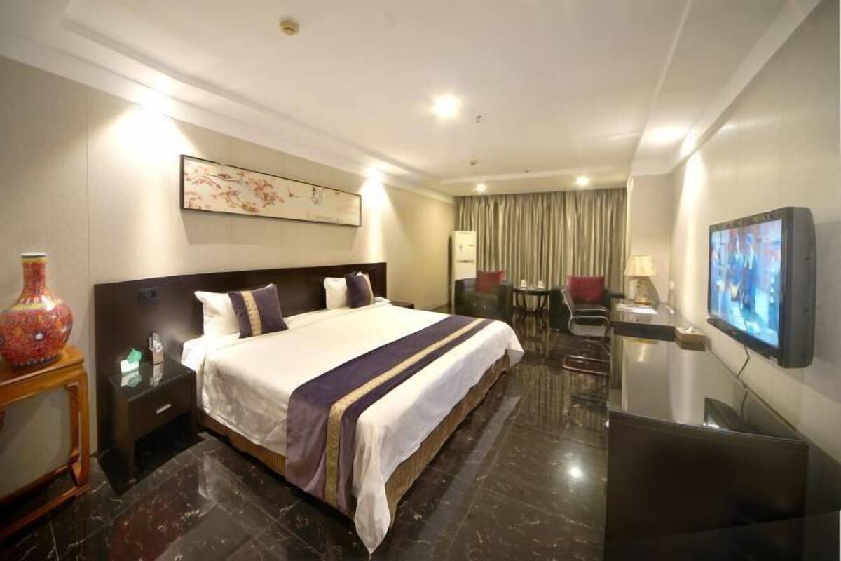 A hotel room with a large bed, a desk with a chair, a television, and a vase on a side table.