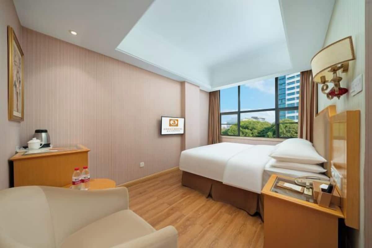 A hotel room with a bed, a desk, a chair, a bedside table, a window with a view, and a framed picture on the wall.