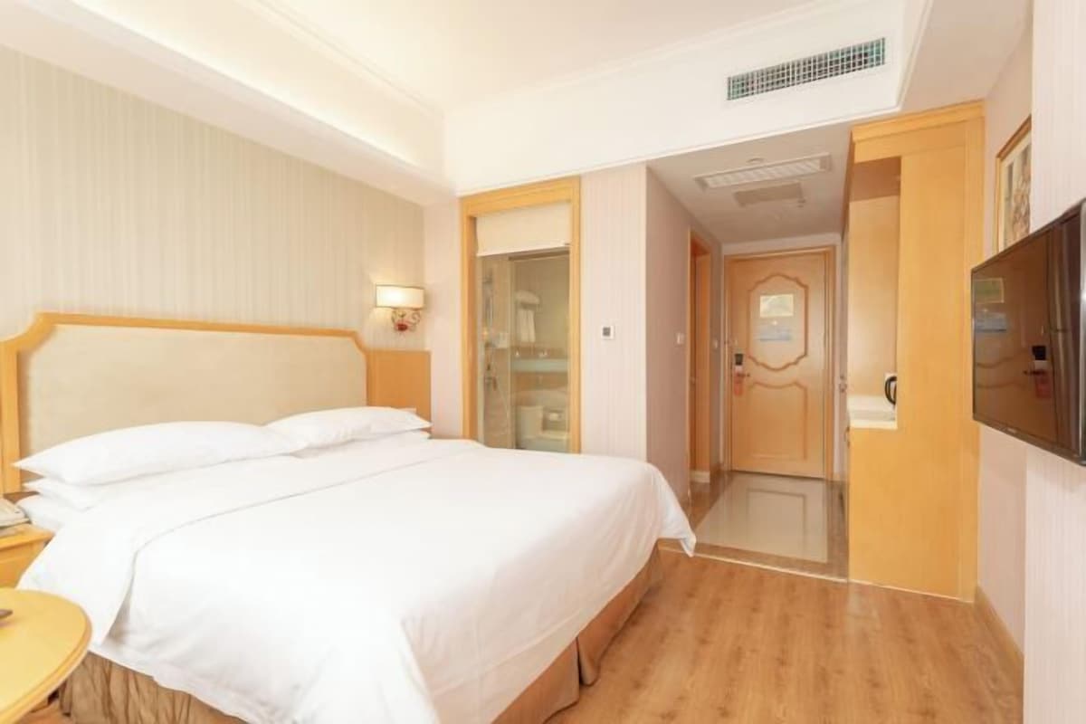 Habitación de hotel con una cama grande, una mesita de noche, un televisor y un baño visible a través de una puerta de vidrio.