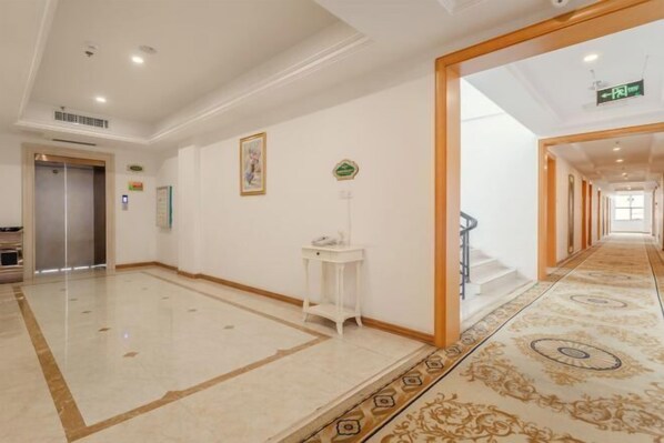 Un pasillo de hotel con suelo de mármol, una alfombra estampada, una mesita blanca pequeña y una escalera que sube a otro piso.