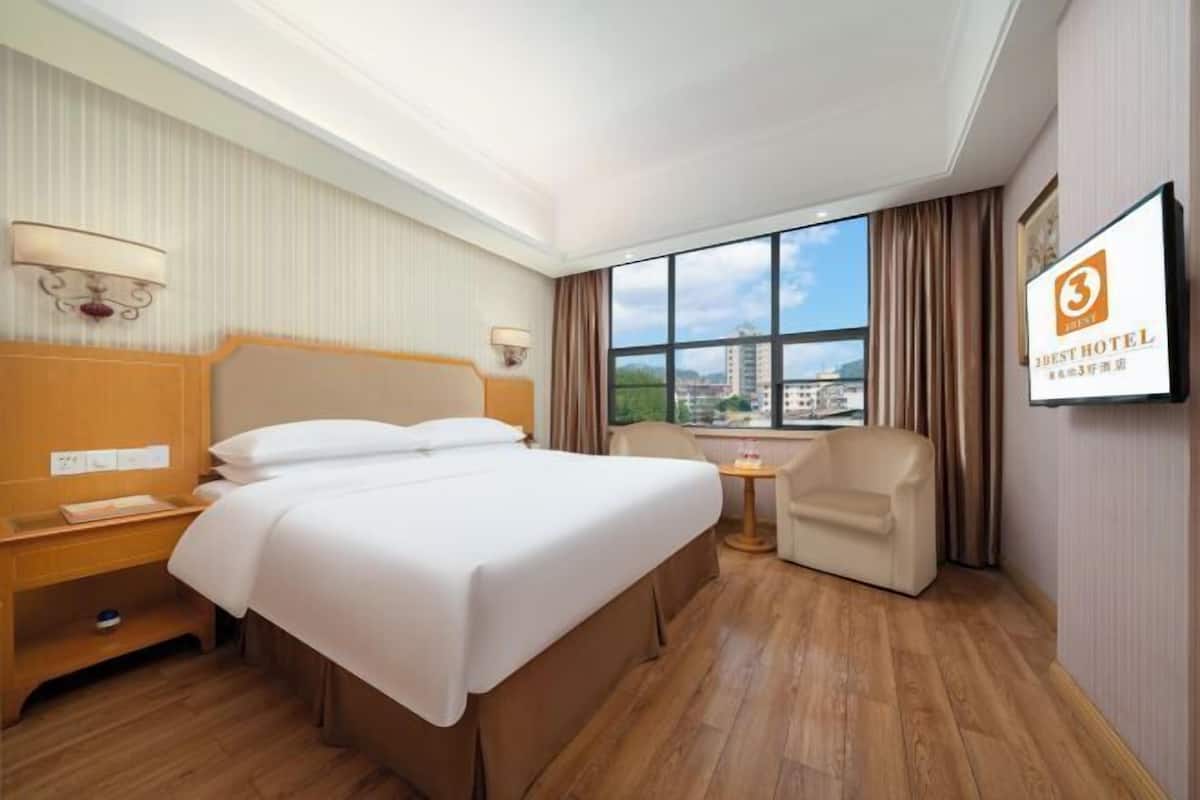 A hotel room with a large bed, a desk, a chair, and a small table. There is a window with a view of buildings outside.