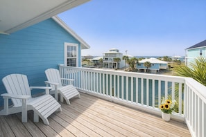 A wooden deck with white Adirondack chairs, a white railing, and a view of the ocean and other buildings.