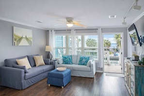A living room with a grey sofa, a blue ottoman, and a white sofa with blue cushions. There is a ceiling fan, a lamp, and a TV mounted on the wall.