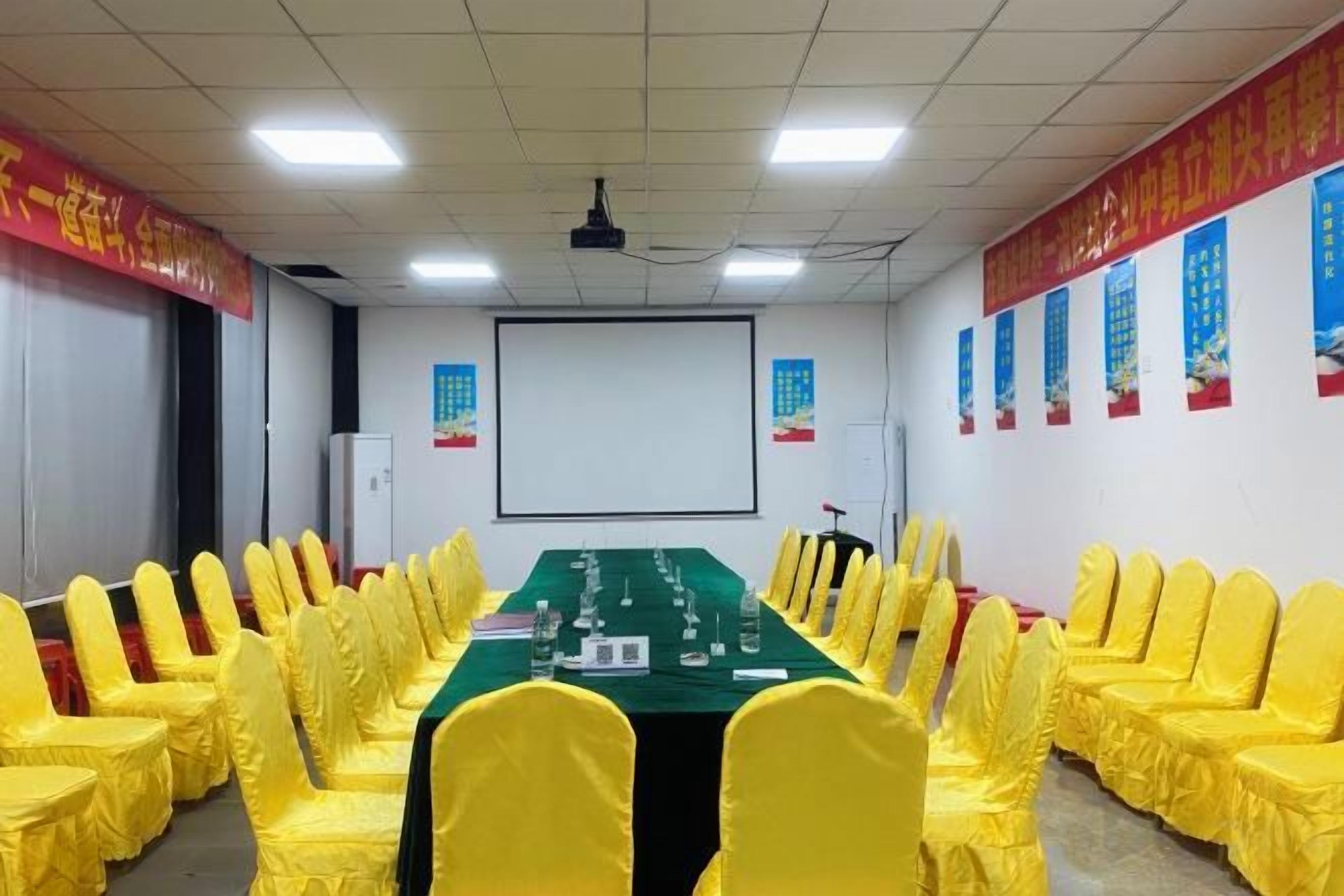 A conference room with a long table, yellow chairs, a projector screen, and red banners on the walls.