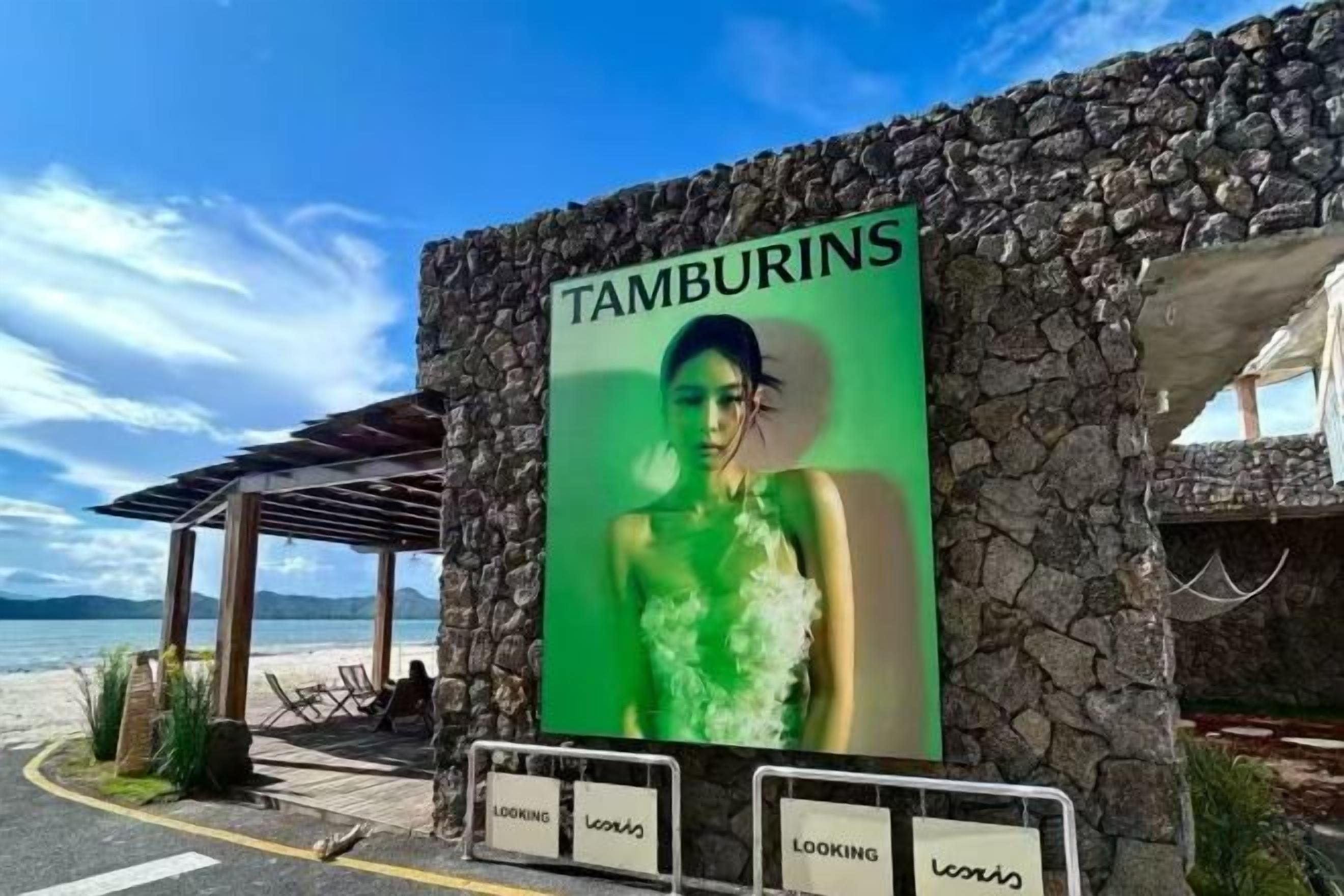 A stone building with a sign reading 'TAMBURINS' and a beach view in the background.