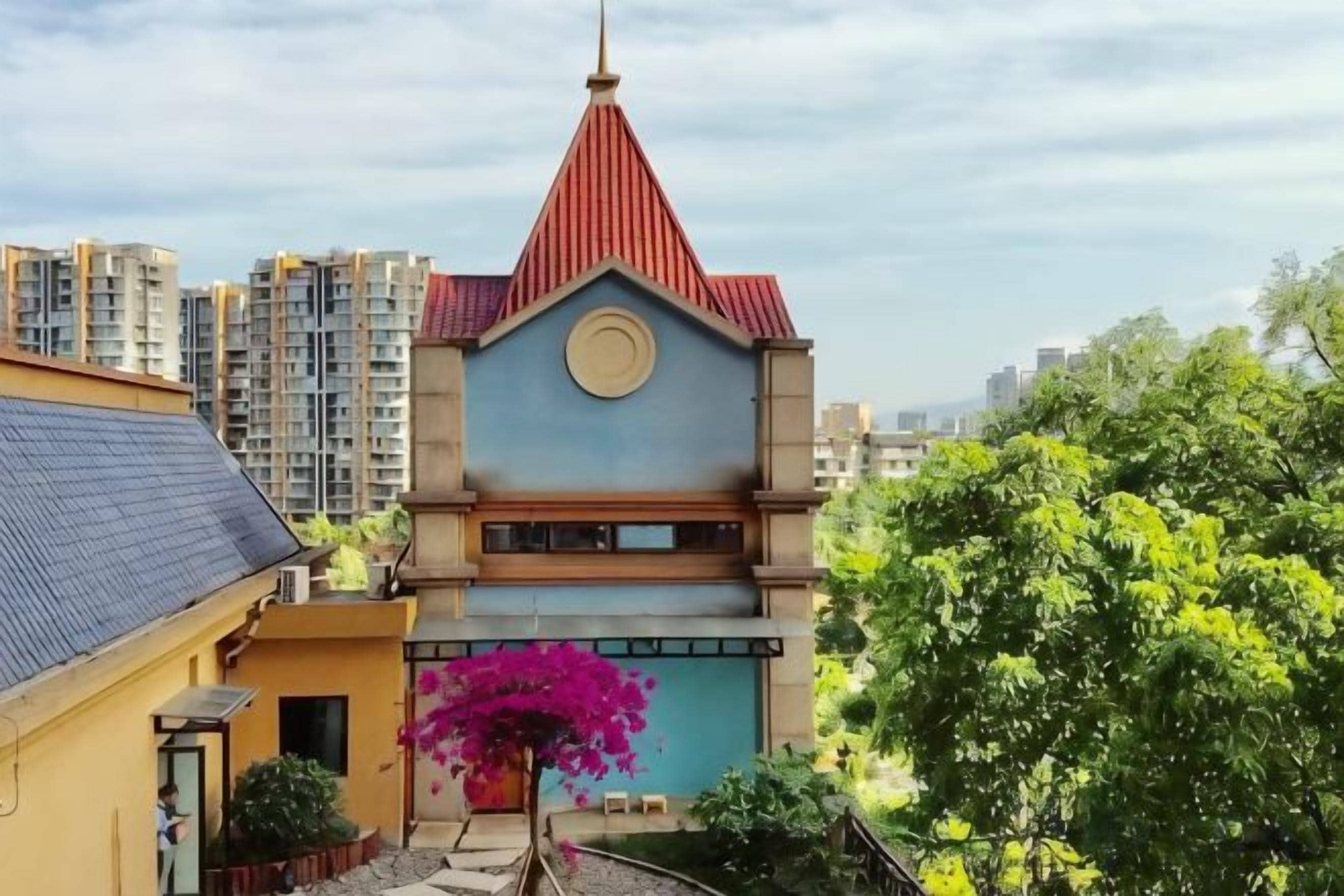 A residential building with a distinctive red roof and a blue facade, surrounded by greenery and other residential structures.