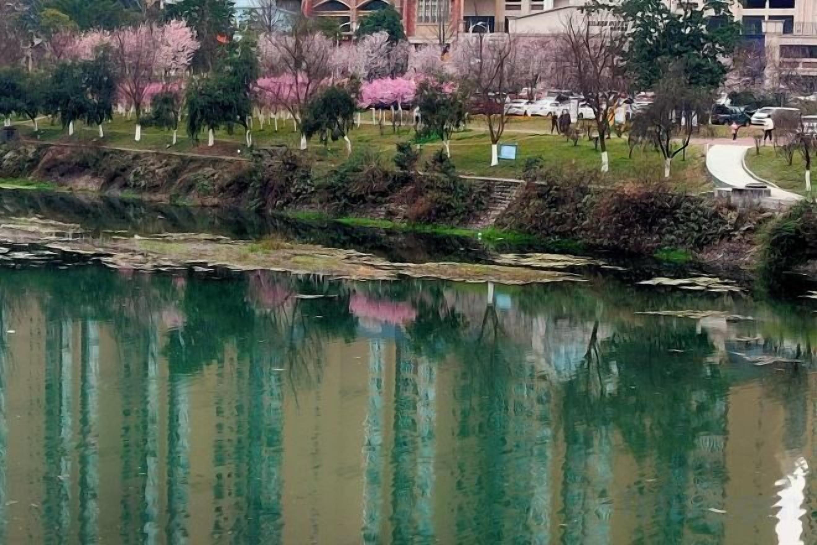 Un río tranquilo con reflejos de árboles y edificios, rodeado de un área ajardinada con senderos.
