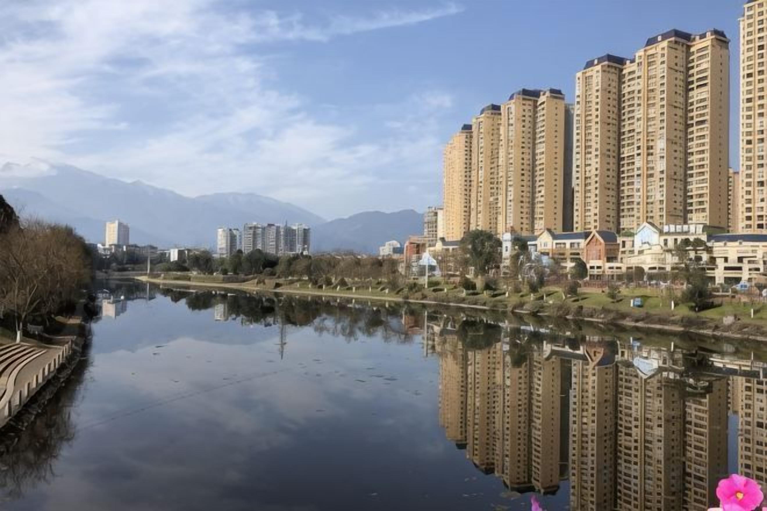 A river reflecting high-rise buildings, a clear sky, and distant mountains.