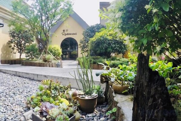A garden area with a pathway leading to a building entrance, surrounded by various potted plants and a large tree.