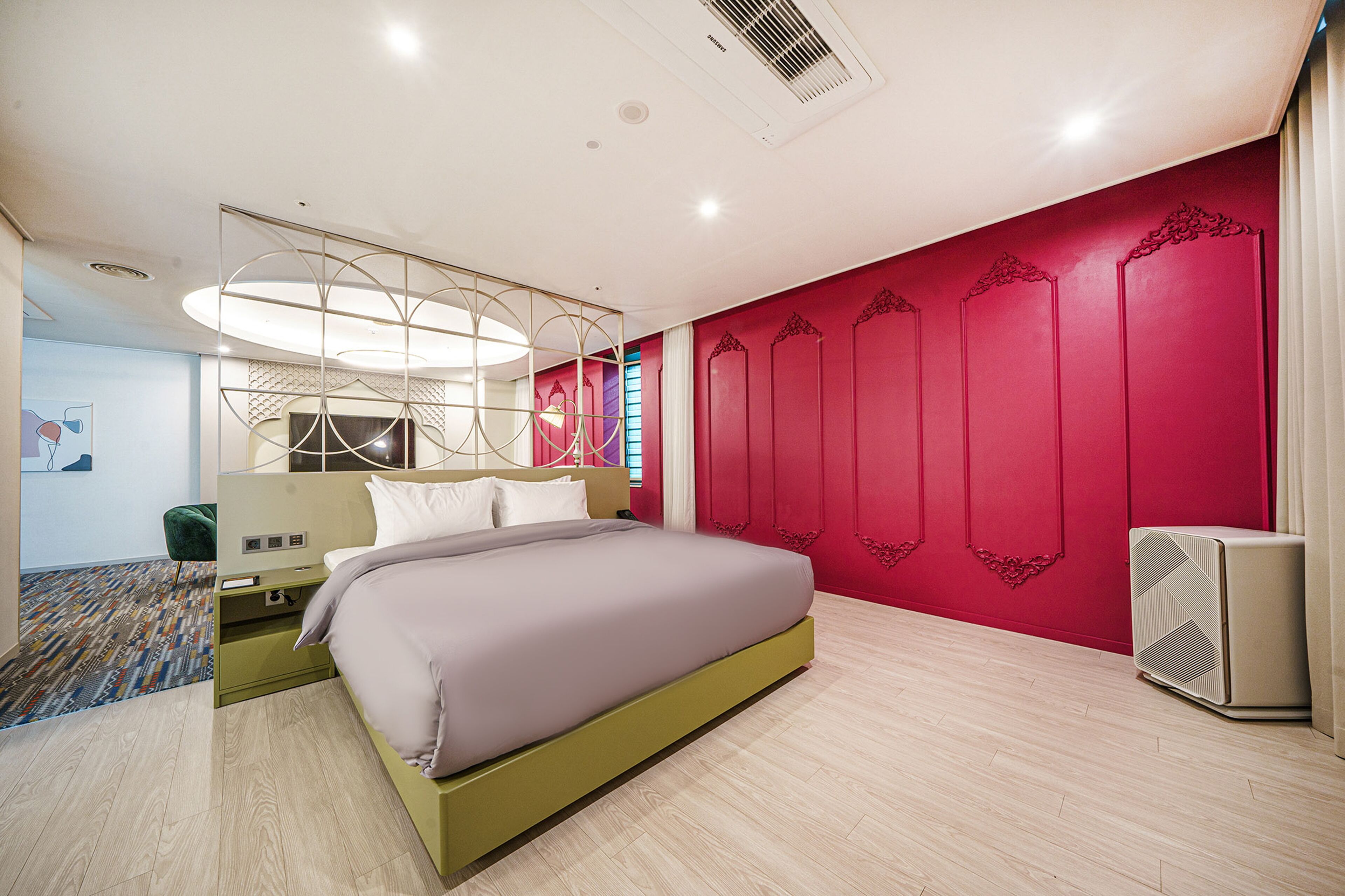 A modern hotel room with a large bed, a bedside table, a nightstand, a wall-mounted air conditioner, and a patterned carpet.