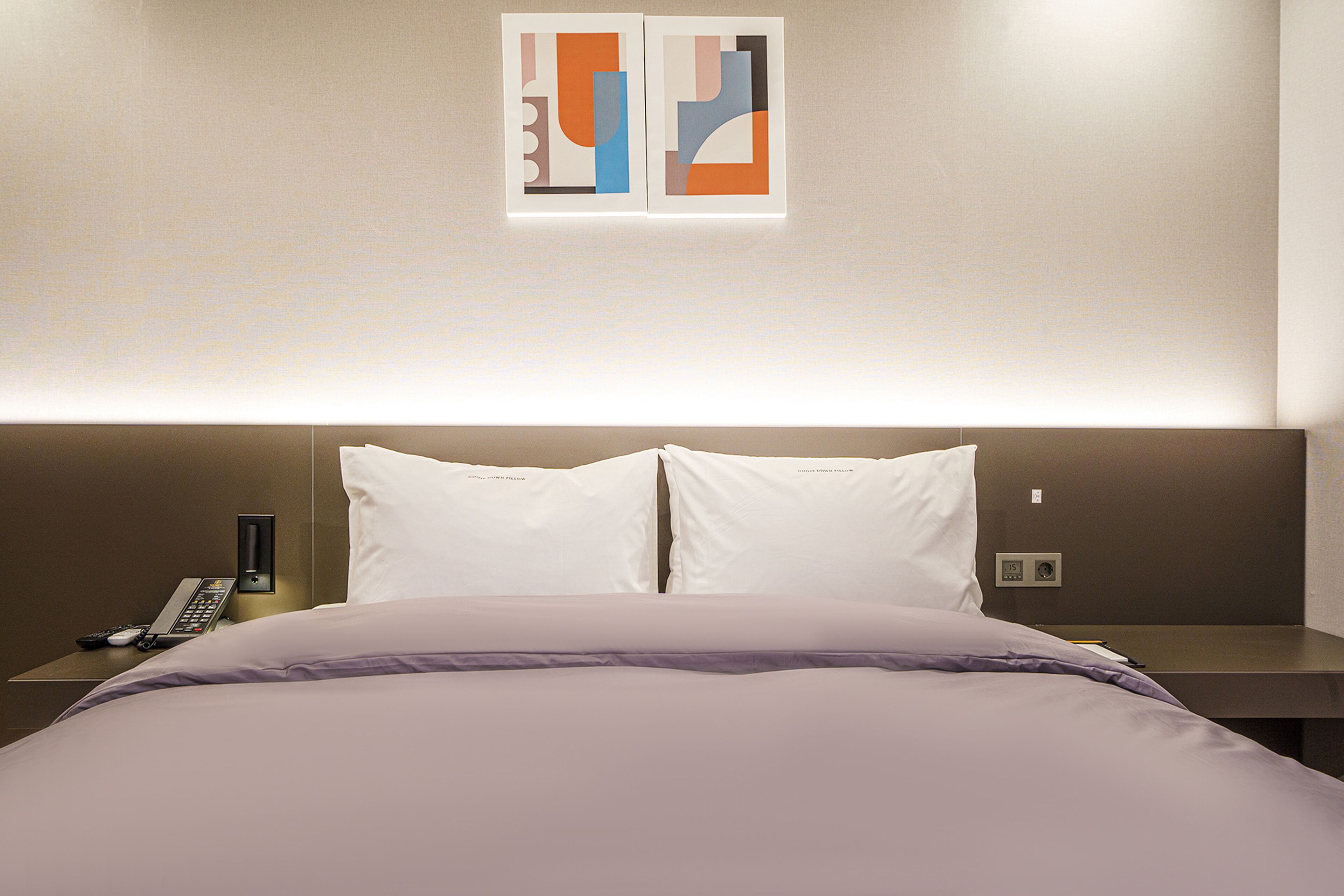 A hotel room with a bed, two pillows, a nightstand with a phone, and a wall-mounted artwork.