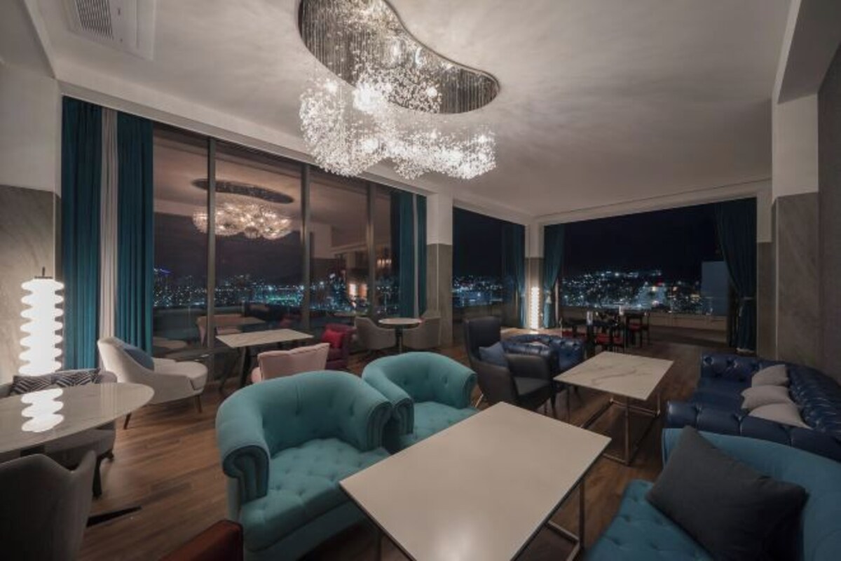 Un espacio lounge moderno con vistas a la ciudad, que incluye una araña, sofás azules y diversas opciones de asientos.