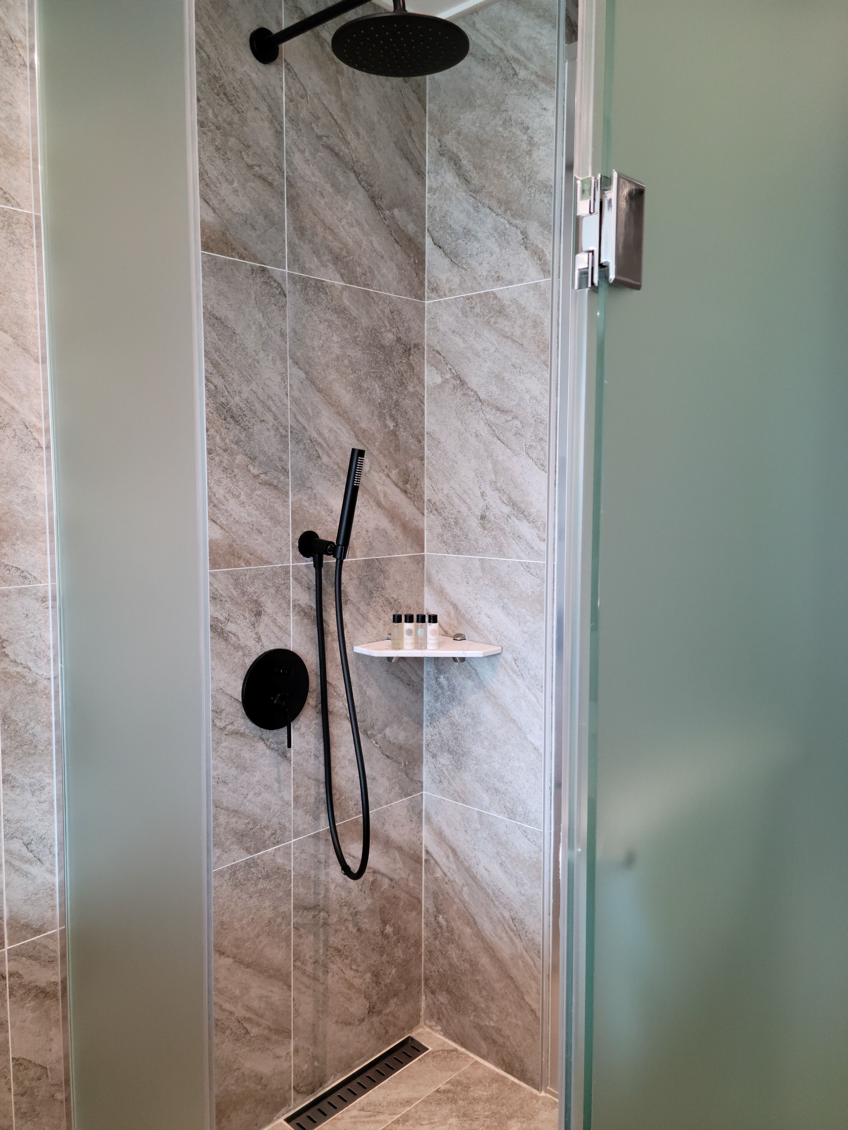 A shower area with a glass door, a handheld showerhead, a wall-mounted showerhead, and a shelf with toiletries.