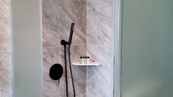 A shower area with a glass door, a handheld showerhead, a wall-mounted showerhead, and a shelf with toiletries.