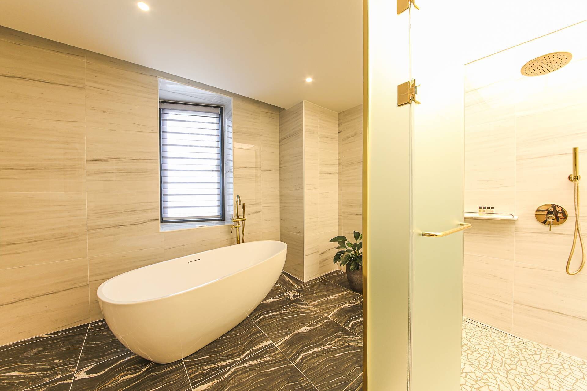 Une salle de bain moderne avec une baignoire sur pieds, une douche dotée d’un robinet doré et une fenêtre avec des stores.