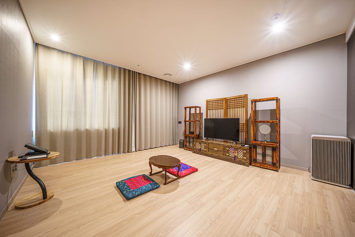 A spacious living room with a large flat-screen TV, a wooden cabinet with a mirror, a small round table, and two colorful rugs.