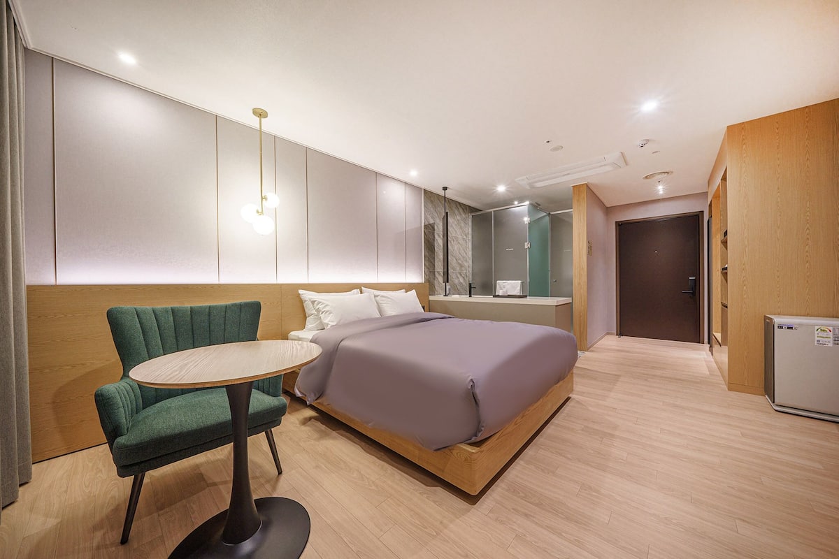 A modern hotel room with a bed, a small round table, a green armchair, and a wooden cabinet.
