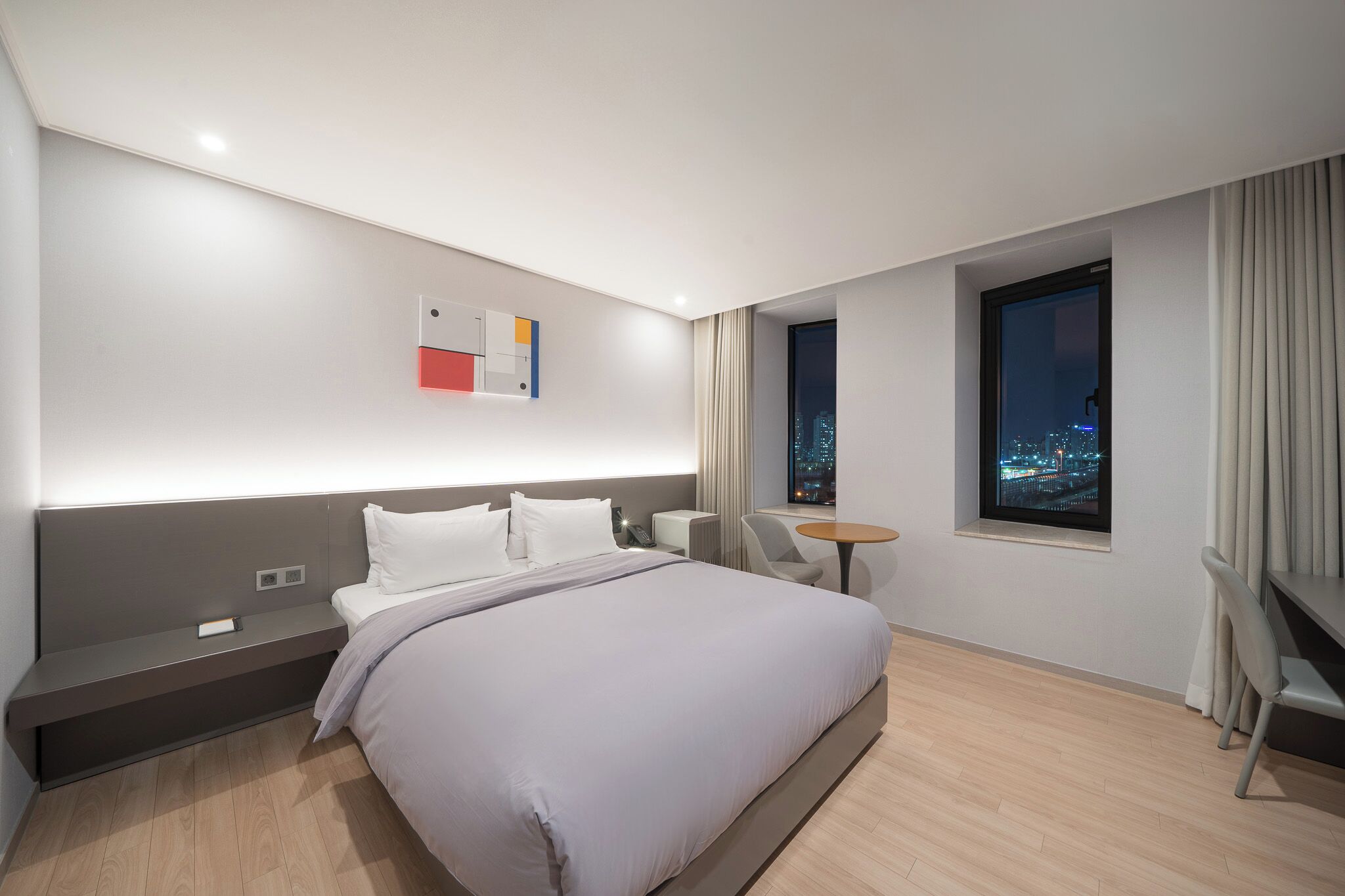 Quarto de hotel moderno com uma cama grande, uma escrivaninha, uma cadeira, uma mesa pequena e uma janela com vista para a cidade.