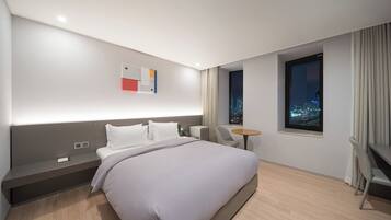 Quarto de hotel moderno com uma cama grande, uma escrivaninha, uma cadeira, uma mesa pequena e uma janela com vista para a cidade.