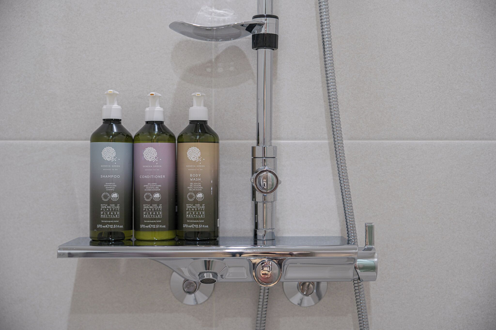 A shower area with a chrome faucet and a handheld showerhead. Two bottles of shampoo and conditioner are placed on a chrome shelf.