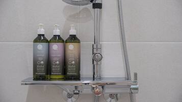 A shower area with a chrome faucet and a handheld showerhead. Two bottles of shampoo and conditioner are placed on a chrome shelf.