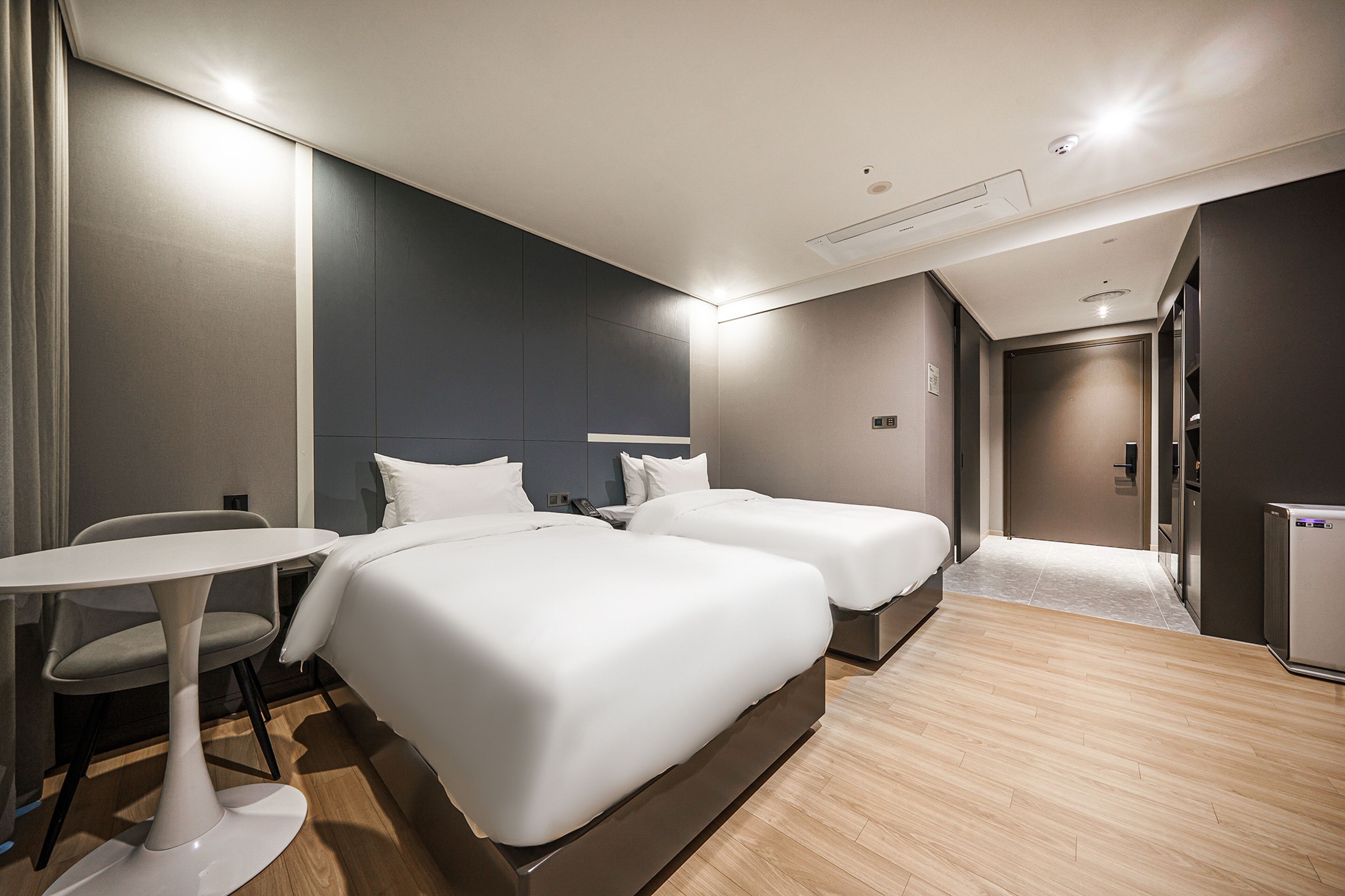 A modern hotel room with two beds, a small table, and a chair.