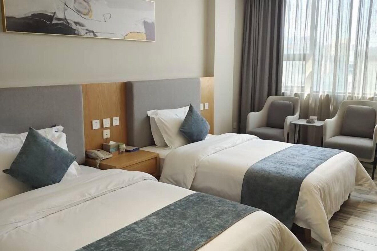 Hotellihuone, jossa on kaksi sänkyä, pieni pöytä, tuoli ja suuri ikkuna, jossa on verhot.