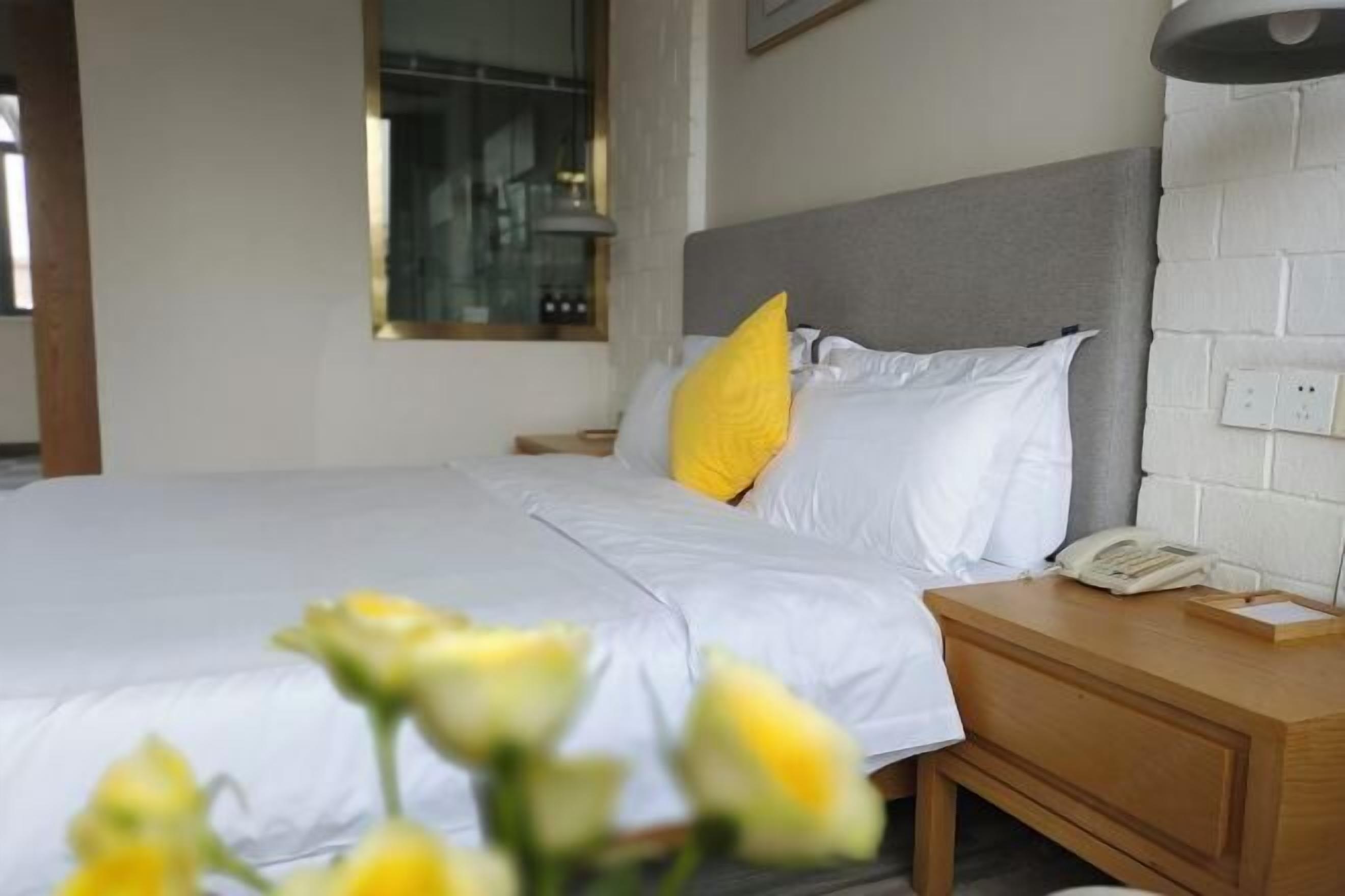 A neatly made bed with white bedding and a single yellow pillow, a bedside table with a lamp, and a wall-mounted light fixture.