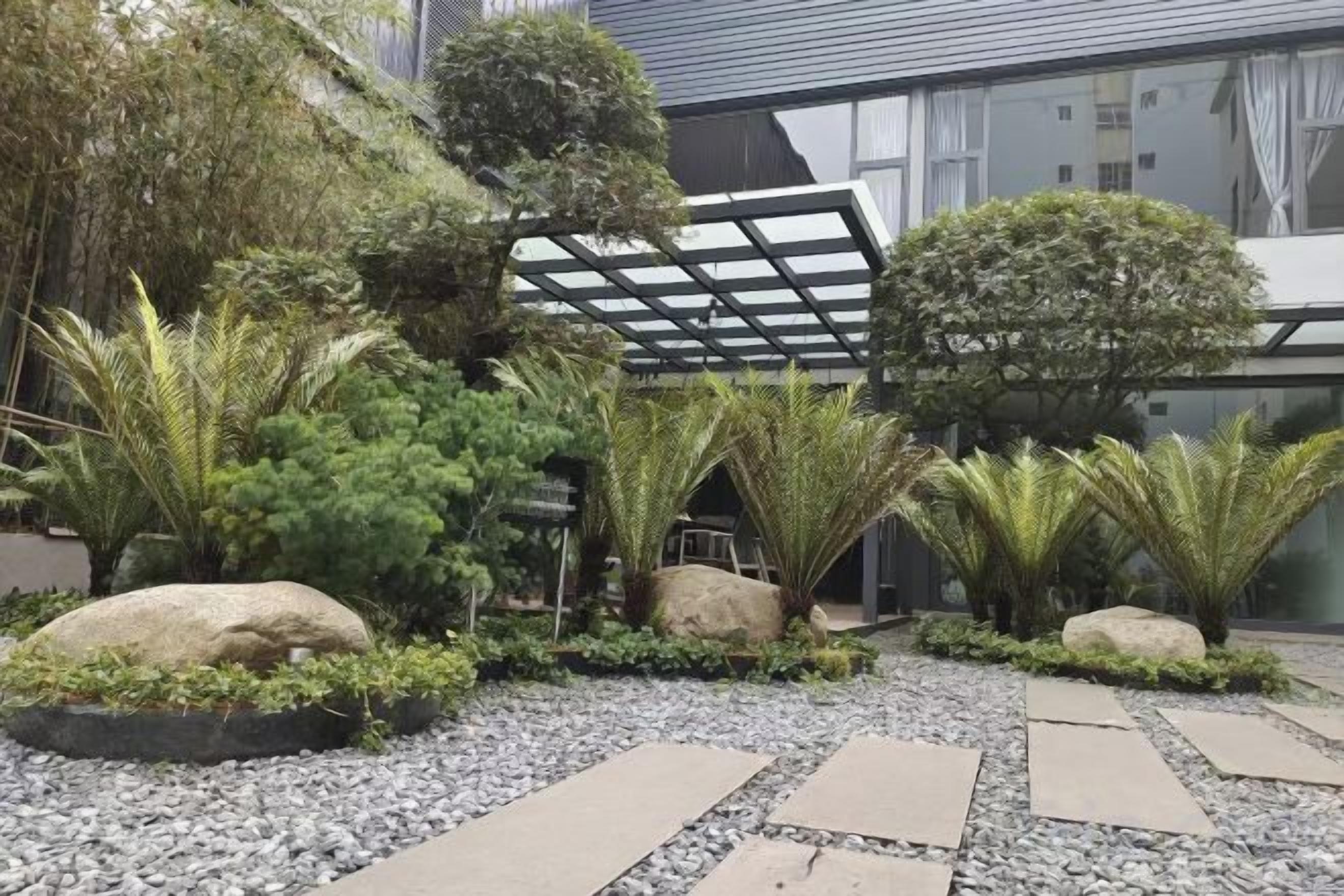 Un jardin aménagé avec différentes plantes et rochers, une structure en forme de pergola et un chemin menant à un bâtiment.