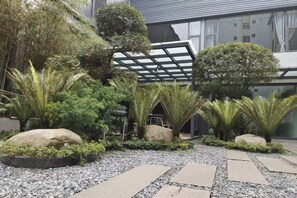 Un jardín ajardinado con diversas plantas y rocas, una estructura de pérgola y un sendero que lleva a un edificio.