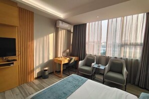 A hotel room with a bed, a desk, two chairs, a TV, and large windows with curtains.