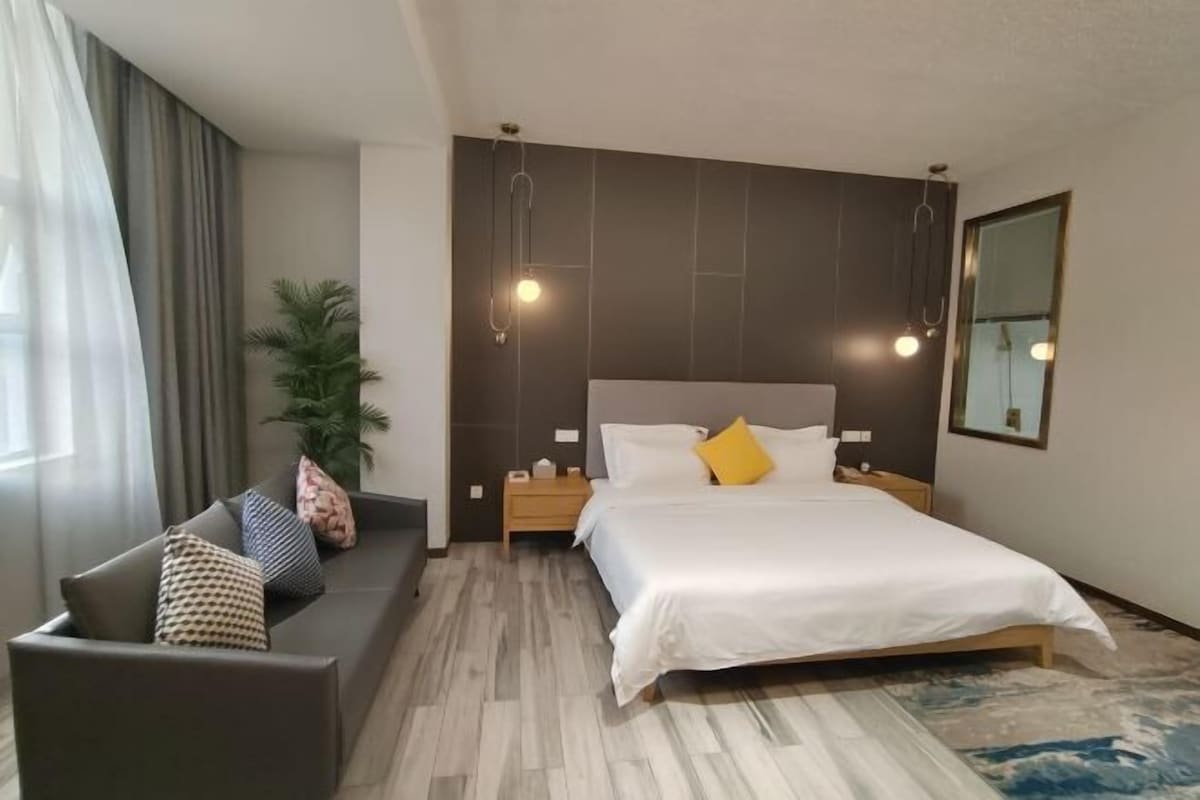 A modern hotel room with a large bed, a sofa, a nightstand, and a potted plant.