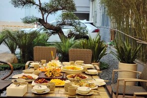 An outdoor dining area with wicker chairs, a table set with various dishes and drinks, surrounded by potted plants and a building in the background.
