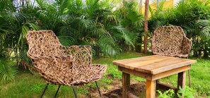 Relax in a serene garden setting with a wicker chair and wooden table amidst lush greenery and palm trees.