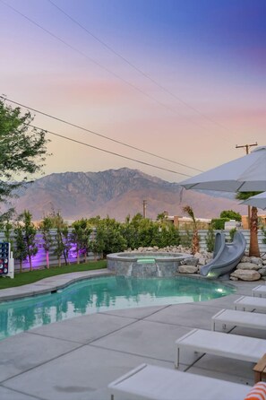 A swimming pool with a slide, lounge chairs, and a view of mountains.