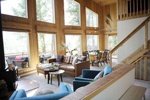 A spacious living area with a large sofa, blue armchairs, and a dining table. There is a wooden staircase leading to an upper level.