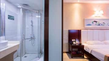 A hotel room with a glass-enclosed shower, a bedside table with toiletries, a bed with white linens, and a wall-mounted light fixture.