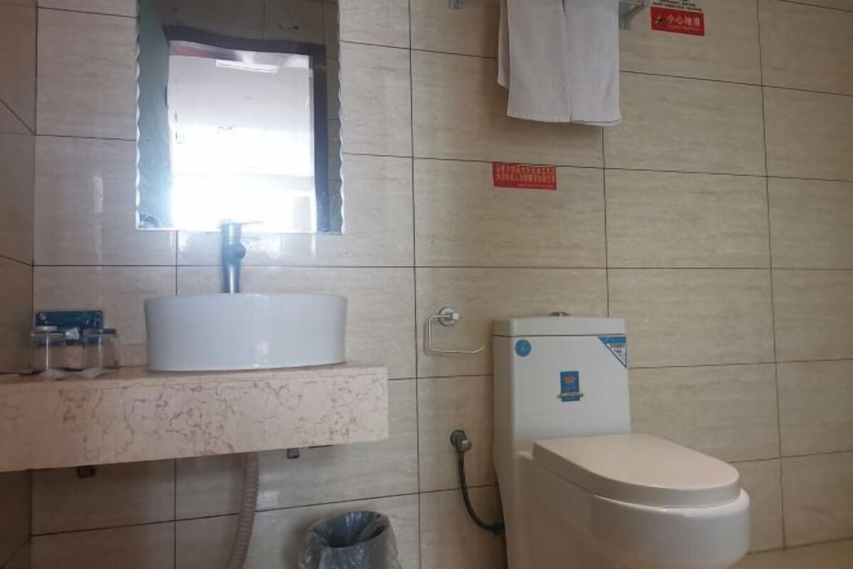 A bathroom with a white sink, mirror, and toilet. There is a trash bin and a towel hanging on the wall.